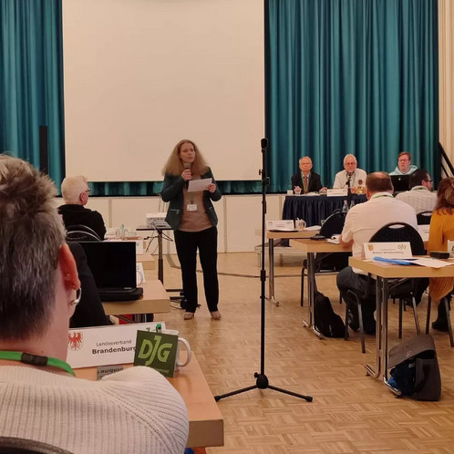 Eine Frau steht in der Mitte eines Seminarraums und spricht vor den anderen Anwesenden in ein Mikrofon