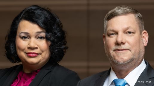 Eine Frau und ein Mann stehen vor einer braunen Wand und blicken in die Kamera