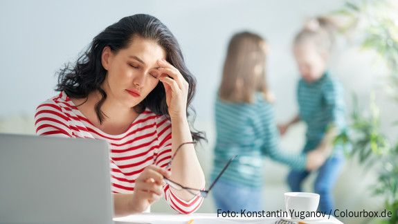 Eine dunkelhaarige Frau arbeitet am Laptop und reibt sich gestresst die Augen, im Hintergrund spielen zwei kleine Mädchen