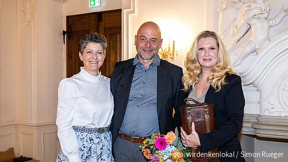 zwei Frauen und ein Mann stehen in einem Konferenzraum und blicken lächelnd in die Kamera